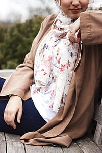 smiling woman wearing white and multicolored floral hijab and brown long-sleeved cardigan holding sunglasses and sitting on wooden bench thumbnail