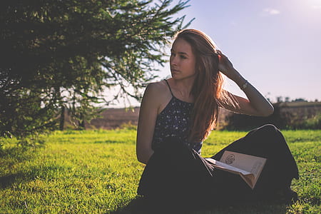 Woman in Black Tank Top and Holding Brown Book Sitting on Grass Under Clear Sunny Sky thumbnail
