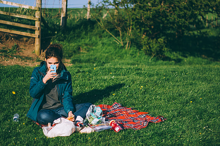 woman in blue jacket holding smartphone sitting on green grass near brown wooden fence during daytime thumbnail