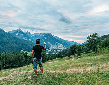 man standing on green grass field holding black Android smartphone near mountain alps thumbnail