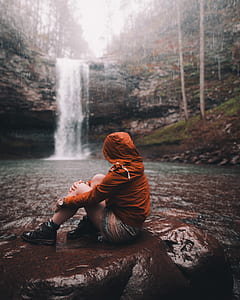 person wearing brown hoodie sitting near waterfalls thumbnail