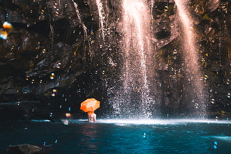 person in water holding orange umbrella under waterfall thumbnail