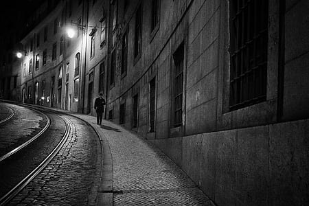 grayscale photo of man walking on street beside concrete buildings thumbnail
