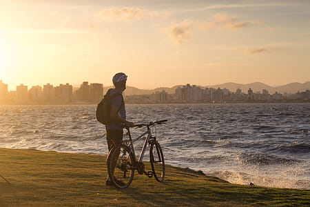 Man With Bicycle Standing on Cliff Front of Water Waves Golden Hour Photography thumbnail