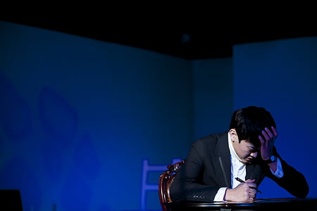 man holding pen while leaning both arms on table and resting his head using left hand behind blue wall thumbnail