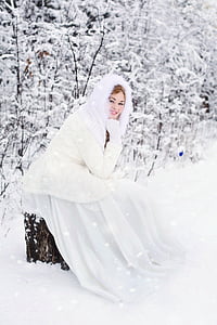 woman wearing white dress and white parka jacket sitting at tree trunk during daytime thumbnail
