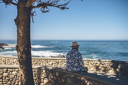 man sitting on concrete bench near tree while facing sea shore thumbnail