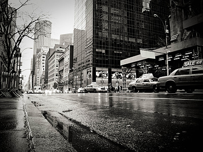low angle grayscale photo of cars passing across the street near city buildings thumbnail