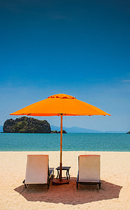 two beach loungers under orange umbrella at seashore under blue calm sky thumbnail