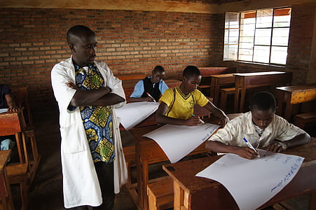 man wearing multicolored shirt and white robe outfit standing next to three men writing on paper in well-lit room during daytime thumbnail
