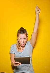 Excited Young Woman Holding An iPad With One Hand Up Against Yellow Wall thumbnail