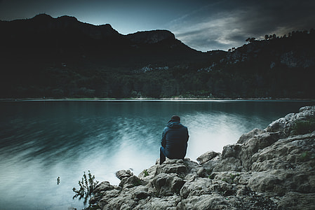 low-light photo of man sitting on cliff of rock formation facing calm body of water thumbnail
