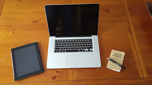 Macbook Pro between black iPad and field notes notebook with black retractable pen on top on brown wooden table thumbnail