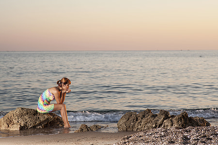 woman in blue and yellow strapless dress sitting on stone near body of water thumbnail