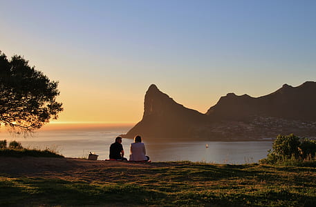 man and woman sitting on green grass field watching mountain and ocean view during golden hour thumbnail
