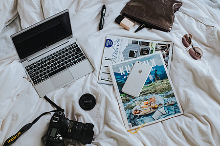 Silver laptop, a camera, magazines and other items on white bed sheets thumbnail