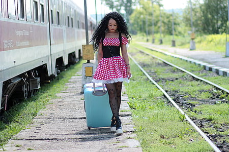 woman in red and white polka-dot dress holding teal luggage beside train during daytime thumbnail