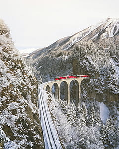 red train passing on rail bridge near mountain covered with snow thumbnail