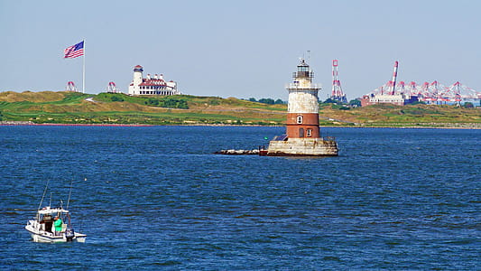 A fisherman fishing from a small boat in New York harbor near the Upper New York Bay Lighthouse with the Bayonne Golf Club in the background. thumbnail