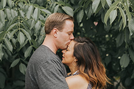 man kissing woman's forehead infront of green leaves thumbnail