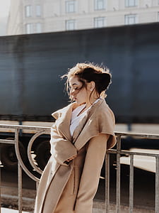 woman wearing brown coat leaning against metal fence during daytime thumbnail