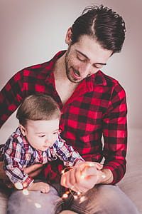 Man Wearing Red and Black Gingham Sports Shirt With Baby Beside Him thumbnail