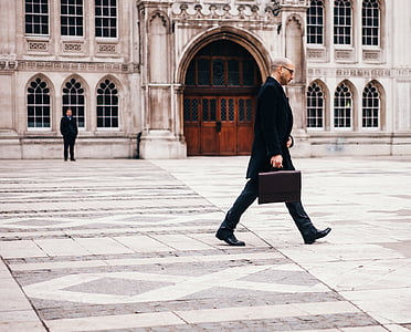 man wearing tuxedo holding brown leather handbag walking on open field near white building thumbnail