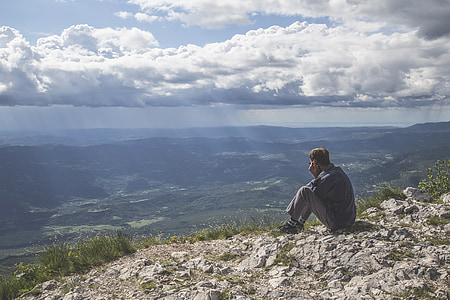 sitting man wearing blue long-sleeved shirt and gray pants in front of green mountain at daytime thumbnail