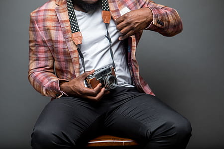 Man Wearing Button-up Shirt Holding Black and Brown Camera While Sitting on Chair thumbnail