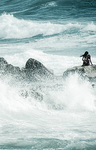 black haired woman sitting on black rock formation surrounded by body of water thumbnail
