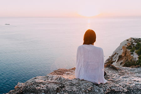 woman wearing white cover-ups sitting on rocky hill with the view of body of water during golden hour thumbnail