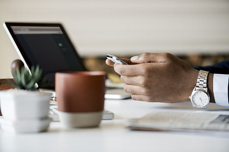person wearing round white analog watch while holding smartphone in front of turned-on laptop computer thumbnail