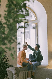 Man Wearing Green Jacket in Front of Man Wearing Orange Zip-up Jacket Sitting on Near the Window Photo thumbnail