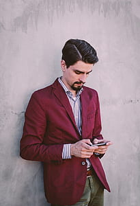 Man Wearing Maroon Blazer Leaning on Gray Concrete Wall While Using His Smartphone thumbnail