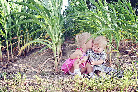 girl wearing pink dress sitting near grass field kissing boy wearing gray shirt and shorts during daytime thumbnail