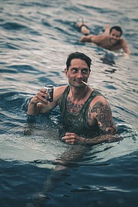 selective focus photography of man in water wearing gray tank top holding can and smoking cigarette thumbnail
