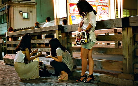 two women squatting and one woman standing beside brown wooden handrail during nighttime thumbnail