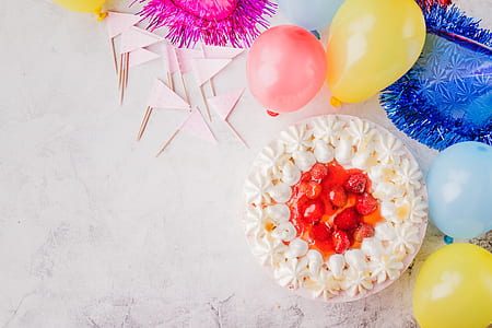 white cake with red sliced fruit beside yellow and blue balloons thumbnail