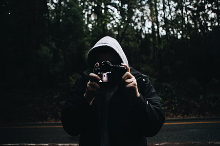 person wearing hoodie holding camera near road and forest thumbnail
