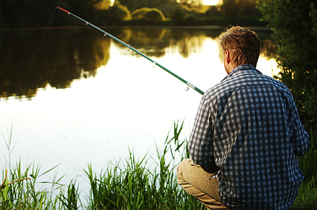 man in blue and white gingham-pattern collard shirt holding green fishing rod while sitting in front body of water thumbnail