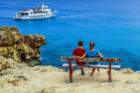 boy and girl seat on brown bench facing on ocean and boat during daytime thumbnail
