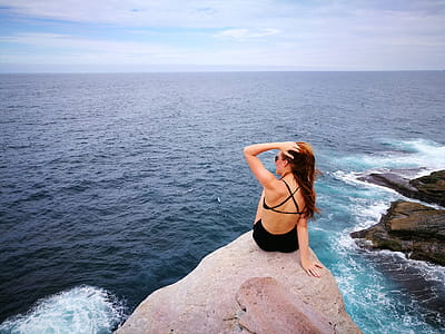 Woman in Black Swimwear Set Sitting on Top of Gray Rocky Cliff Facing Body of Water thumbnail