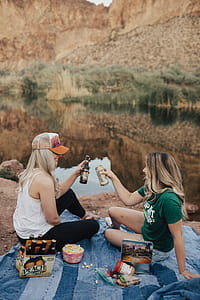 Two Women Holding Beer While Sitting on Rock Near Body of Water thumbnail