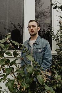 man standing near building window and plant while wearing blue denim jacket during daytime thumbnail