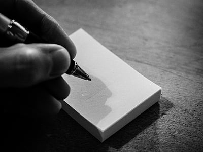 grayscale photography of person holding pen beside note wooden surface thumbnail