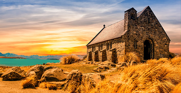 landscape photography of brown and black brick church in the middle of brown grass near body of water during golden hour thumbnail