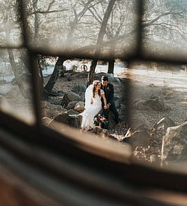 couple sitting on rock while taking photo under tall trees at daytime thumbnail