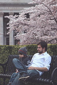 man wearing white button-up long-sleeved shirt sitting on black bench beside toddler wearing gray knitted hat thumbnail