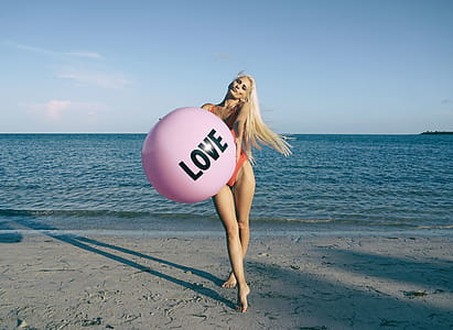 Woman Wearing Orange Swimsuit While Holding Pink Inflatable Beside Body of Water thumbnail