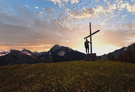silhouetto photo of man standing and holding cross in front of mountain during sunset thumbnail